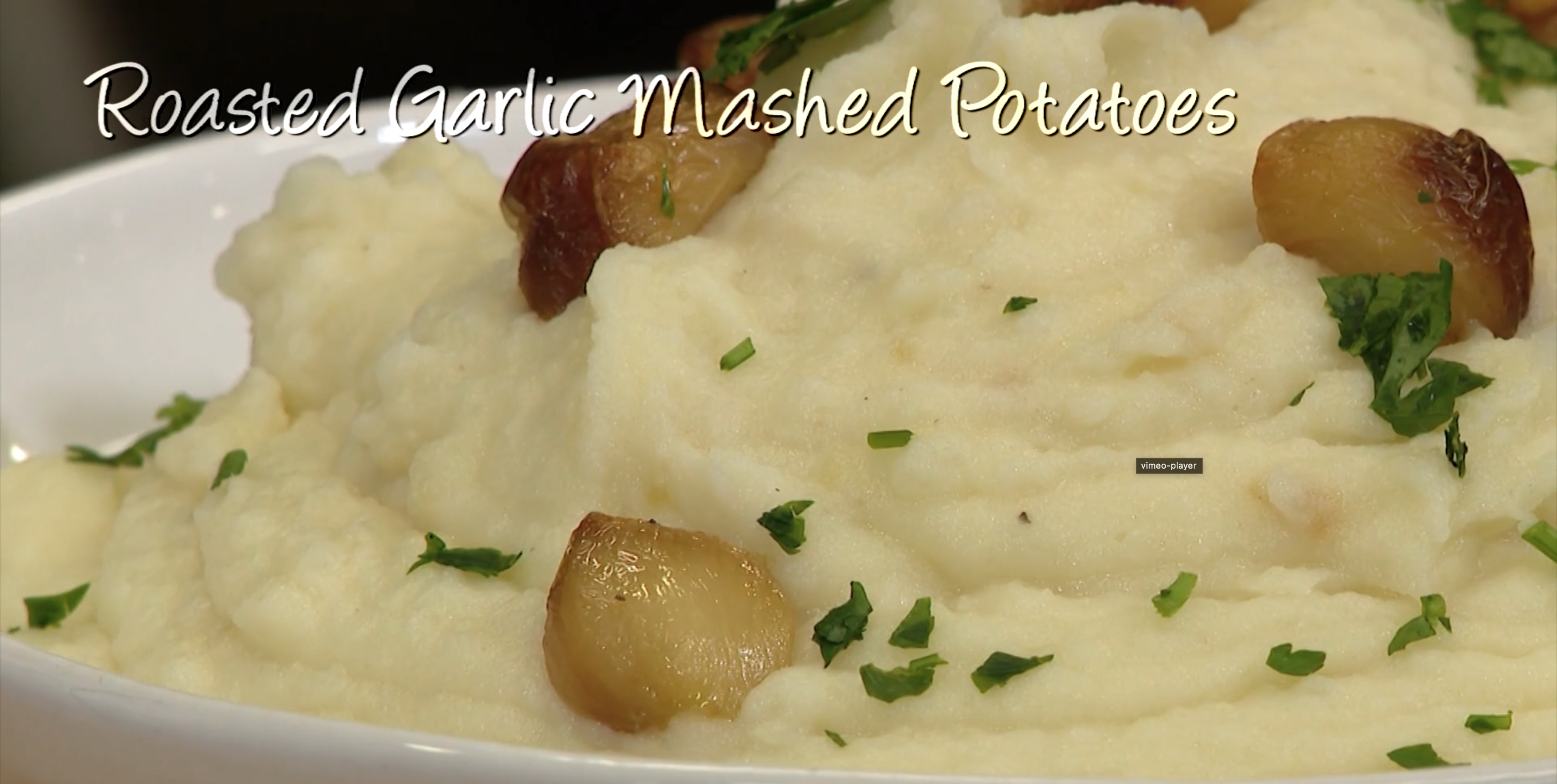 Close up shot of mashed potatoes in a white bowl garnished with garlic and parsley with the text "Roasted Garlic Mashed Potatoes"