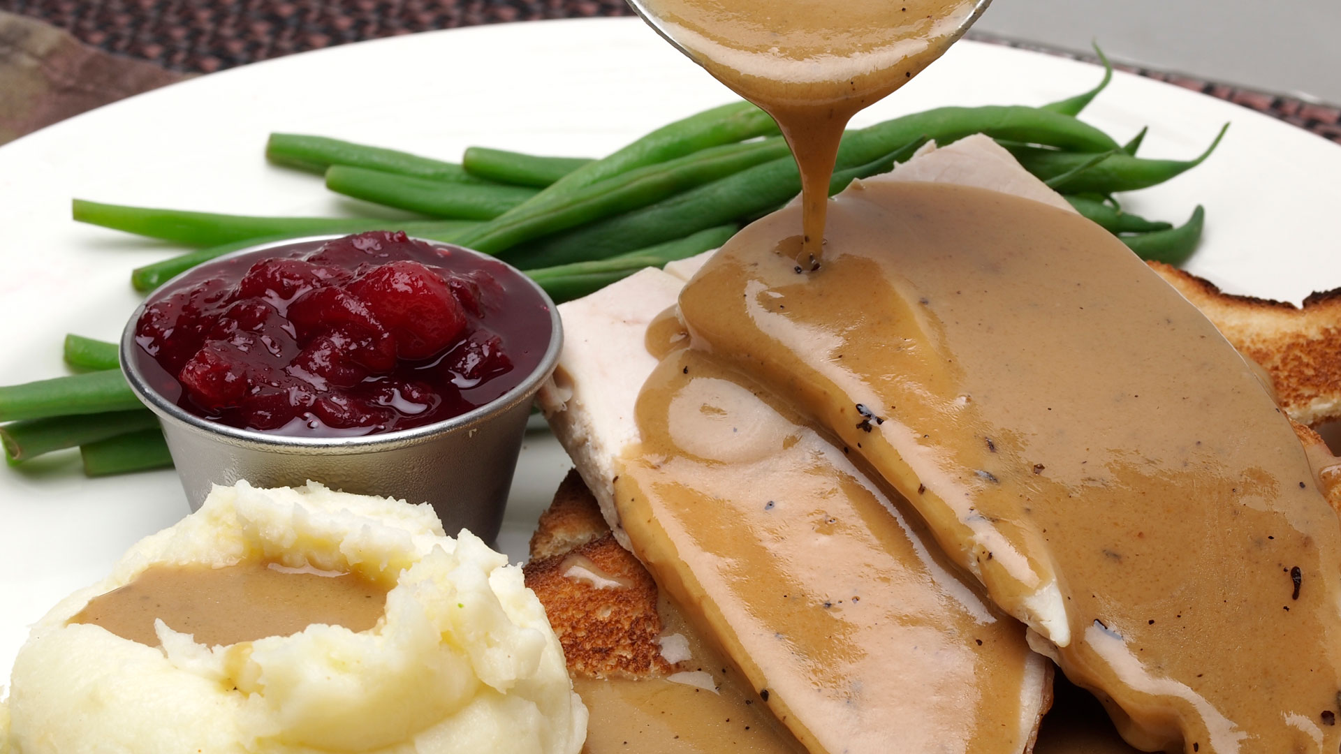 Turkey gravy pouring on a plat bof turkey, mashed potatoes and green beans.